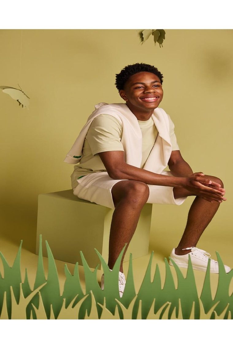 A teenage BE.Kids model in a monochrome set, seated on a cube against a playful paper grass-and-butterfly backdrop