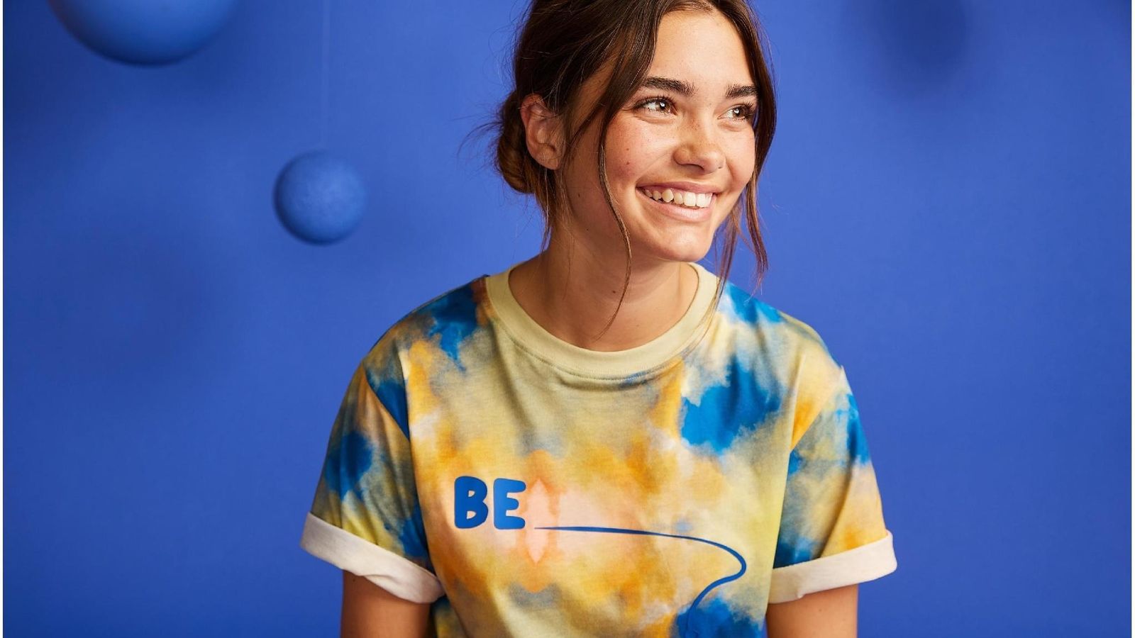 A BE.Kids model smiling in the tie-dye tee against a deep-blue backdrop