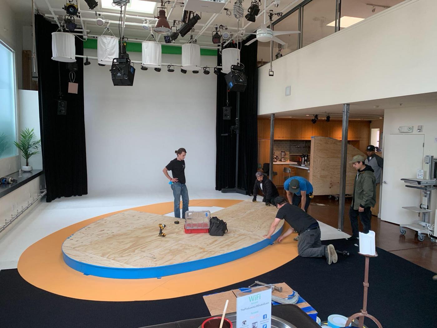 Crew members kneeling to fit the blue-and-orange trim around a plywood oval stage deck under a studio lighting grid