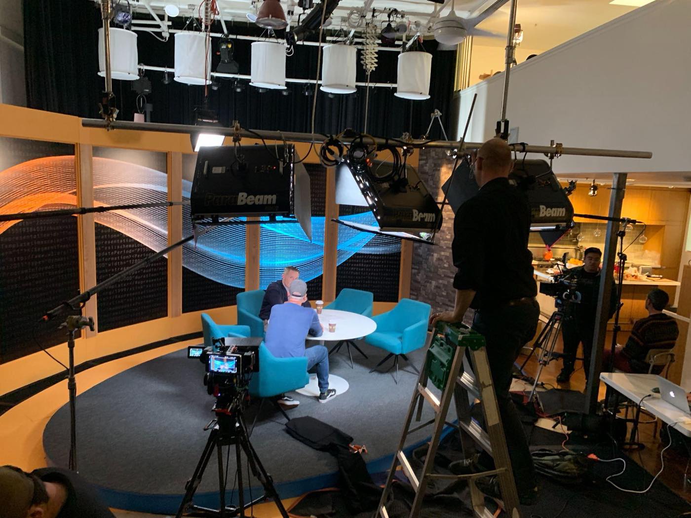 Wide view of the lit set — ParaBeam lights and a boom over the oval stage where the host and crew work at the white table