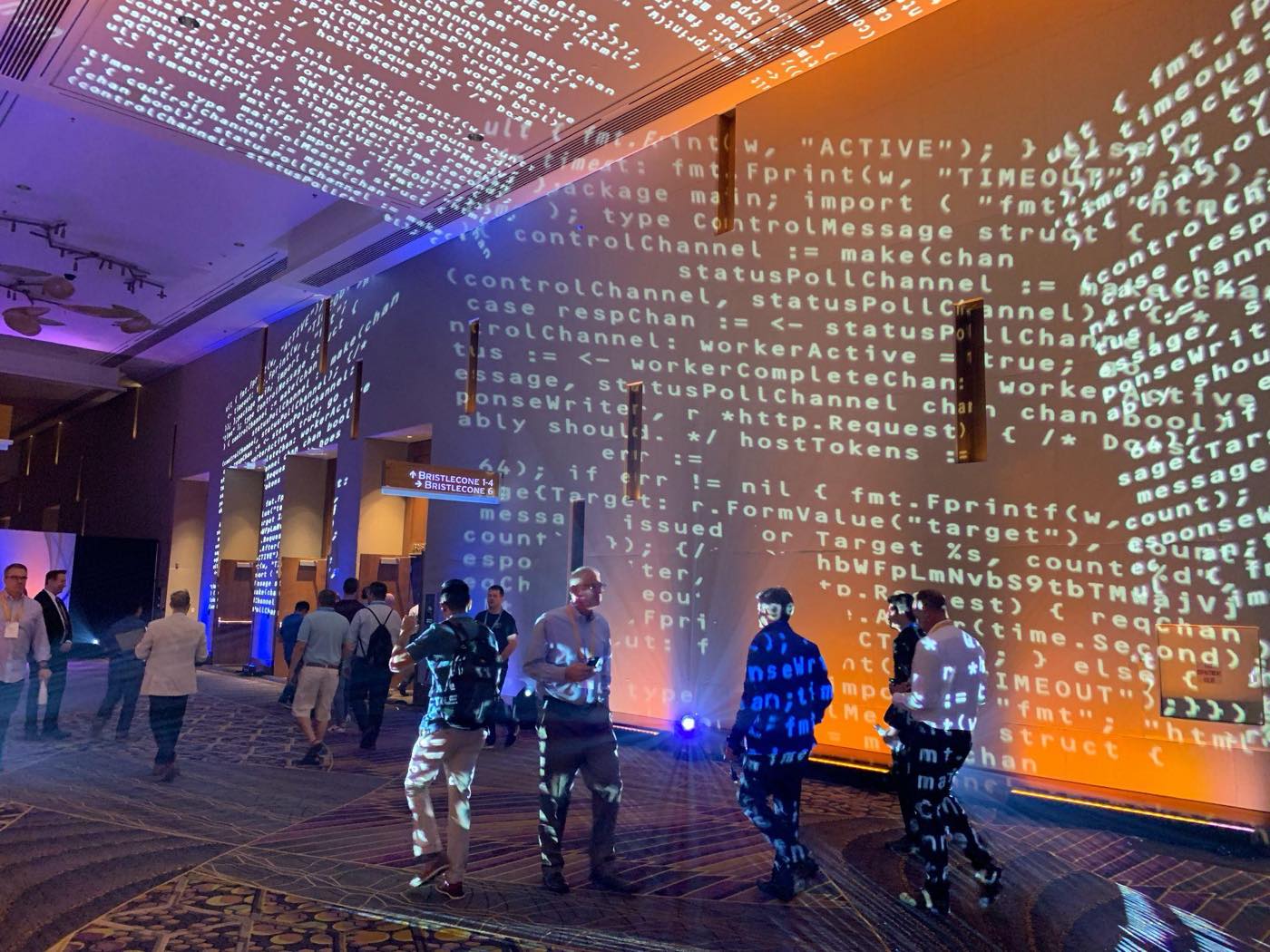 Attendees walking through a conference corridor while lines of Go source code are projected over the walls, ceiling, and across them