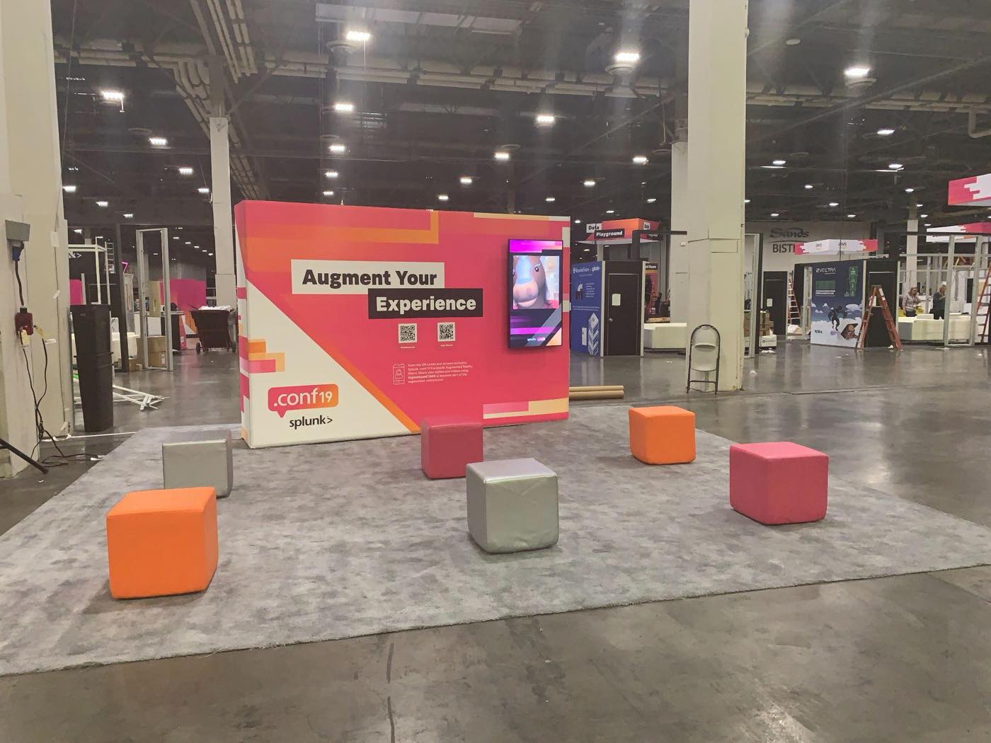 The .conf19 AR wall on the show floor — a pink and orange branded wall reading “Augment Your Experience” with QR codes and a vertical screen, surrounded by coloured seating cubes