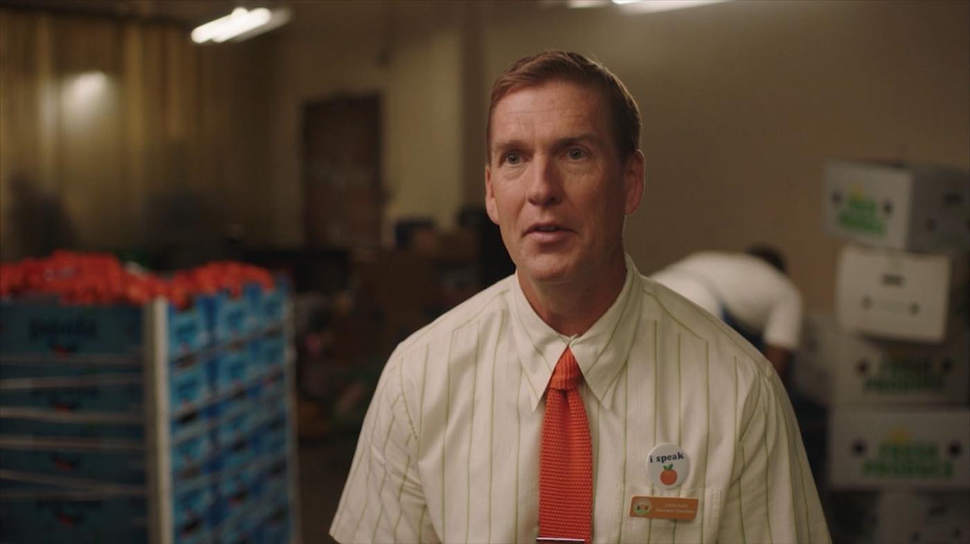 John Birk in a supermarket backroom in a pinstriped shirt and orange tie, wearing an “i speak” button, with pallets of Peelz mandarins behind him