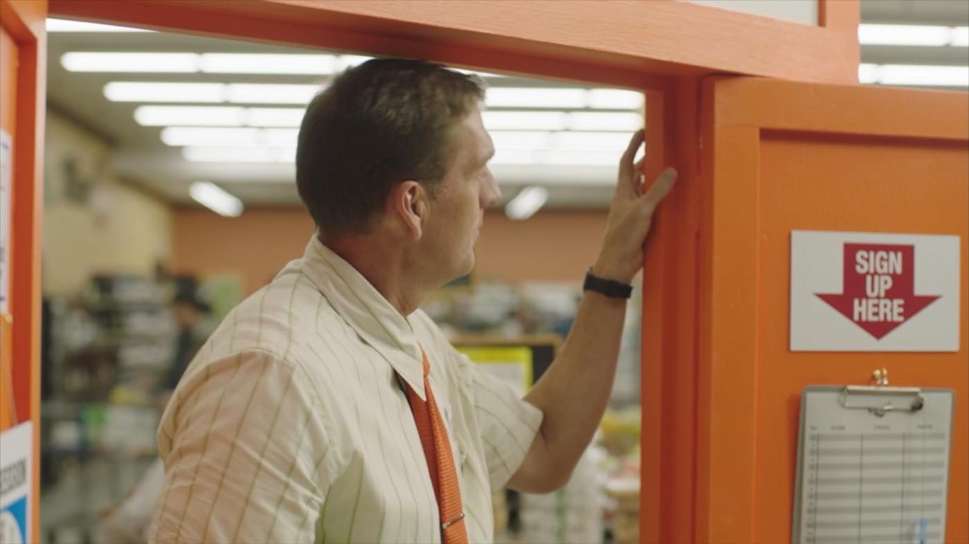 John Birk reaching up to an orange sign-up board marked “Sign Up Here” on the built exterior set at the supermarket