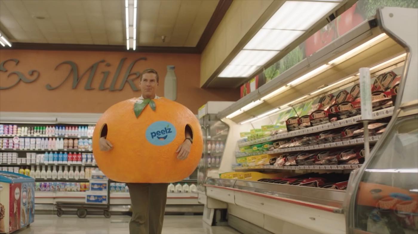 John Birk walking a supermarket dairy aisle in a full mandarin-orange costume with the Peelz logo across the front